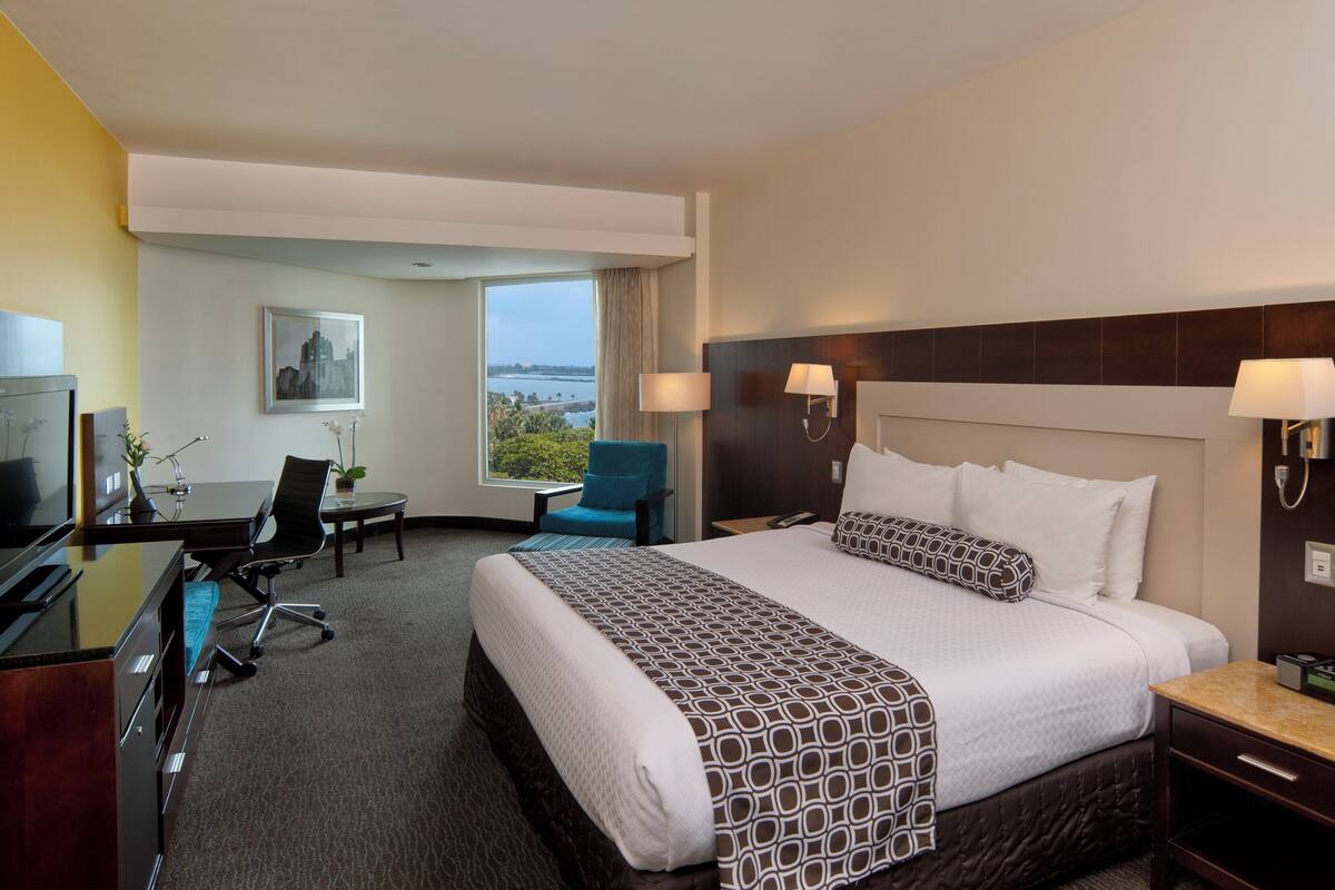 Une chambre d’hôtel avec un grand lit, un bureau avec un ordinateur, une chaise, une télévision et une fenêtre donnant sur l’océan.