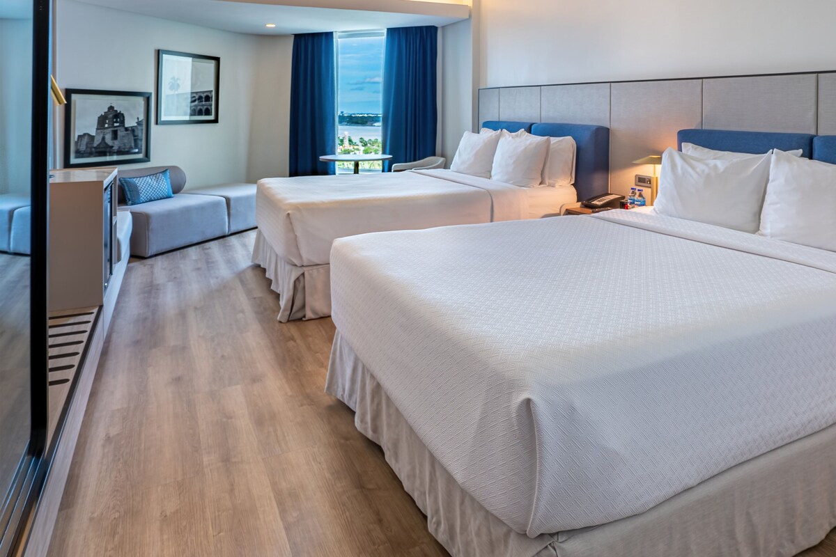 Ein Hotelzimmer mit zwei Betten, einem Sofa, einem Tisch und Blick auf die Stadt.