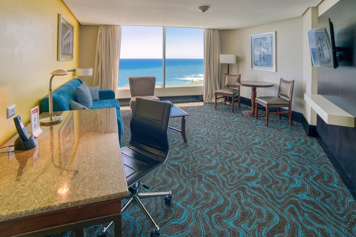 Ein Hotelzimmer mit großem Fenster und Meerblick, einer Sitzecke mit Tisch und Stühlen sowie einer Minibar.