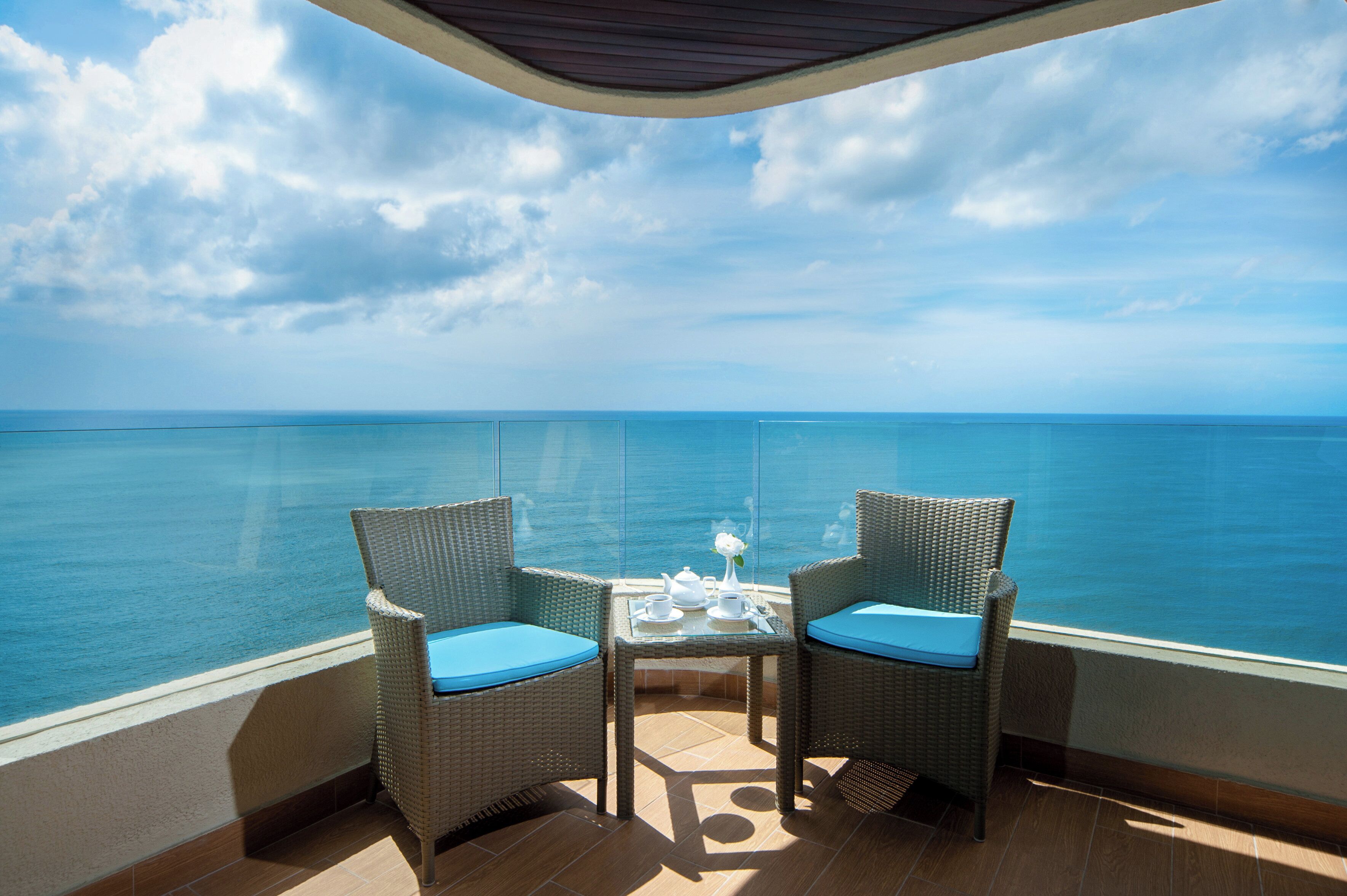 Een balkon met twee rieten stoelen, een klein tafeltje met een theepot, kopjes en een vaas met een bloem, met uitzicht op een heldere blauwe zee.