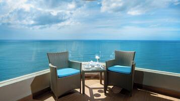 Een balkon met twee rieten stoelen, een klein tafeltje met een theepot, kopjes en een vaas met een bloem, met uitzicht op een heldere blauwe zee.