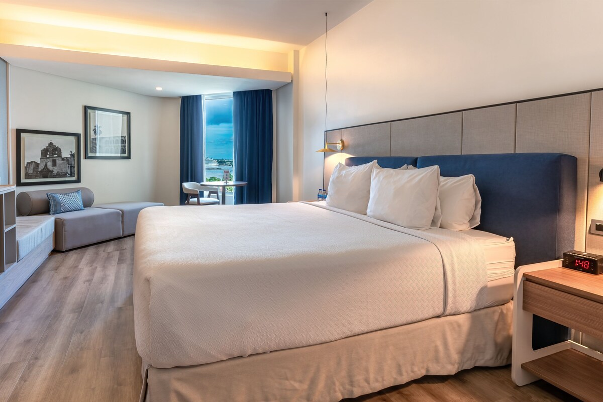 Ein Hotelzimmer mit einem großen Bett, einem Schreibtisch, einem Sessel und Blick auf die Stadt.