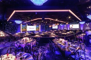 A banquet hall with round tables set for dining, a stage with a large screen, and a chandelier. - The Beverly Hilton (Beverly Hills)