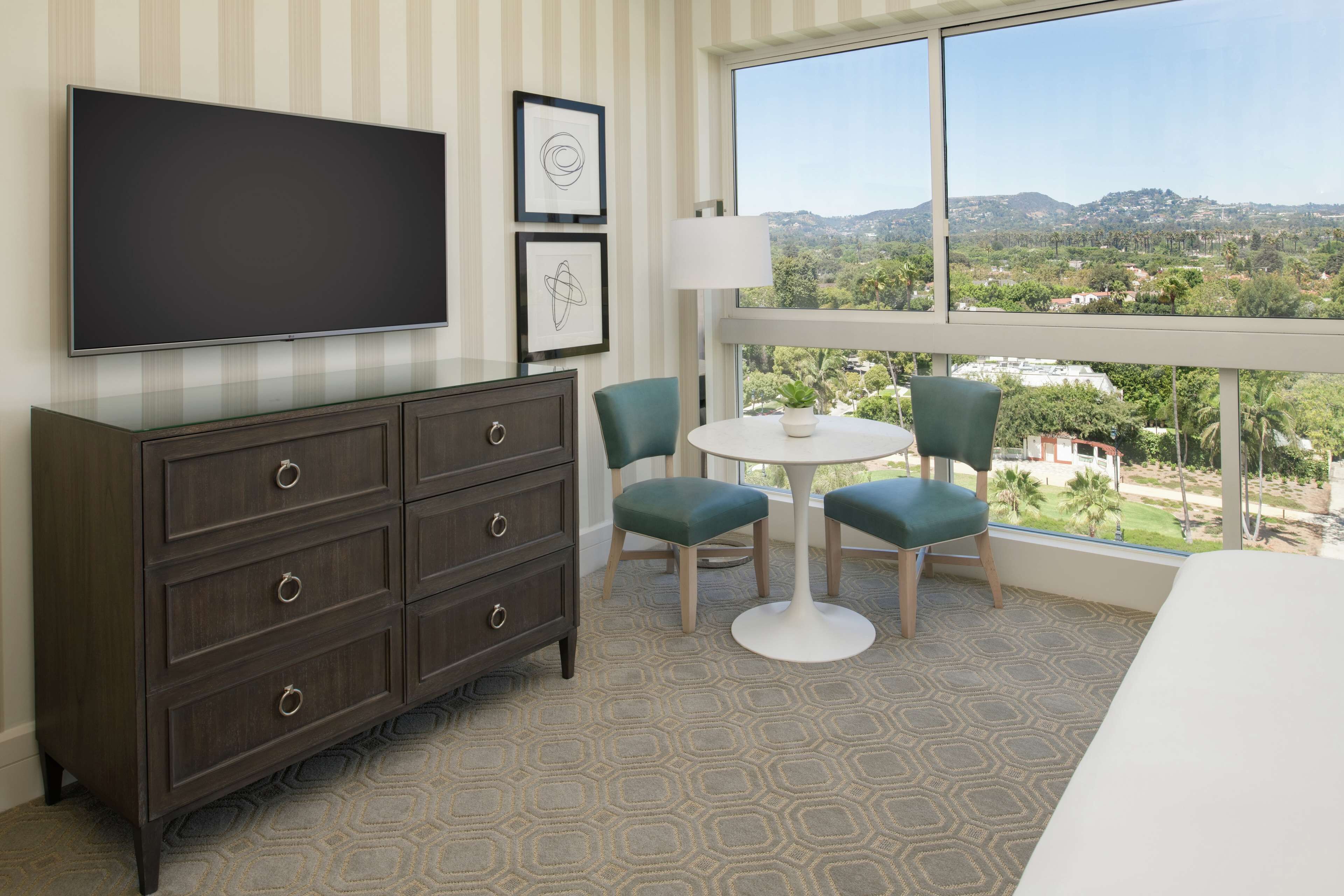 Habitación de hotel con televisor grande, cómoda, dos sillas, una mesita y vistas a un paisaje urbano a través de un ventanal amplio.