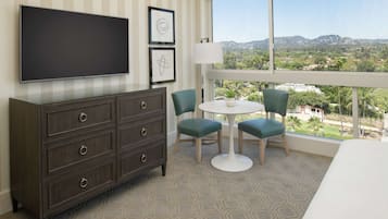 Habitación de hotel con televisor grande, cómoda, dos sillas, una mesita y vistas a un paisaje urbano a través de un ventanal amplio.