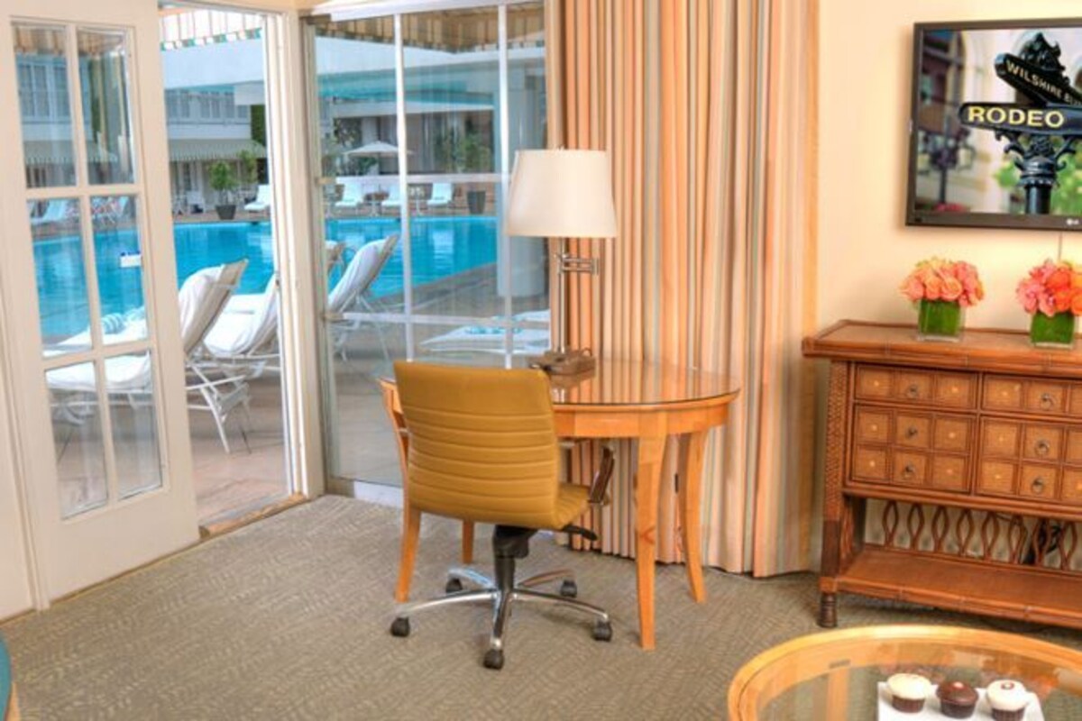 A hotel room with a glass door leading to a pool area, a wooden desk with a lamp, a chair, and a small table with items on it.