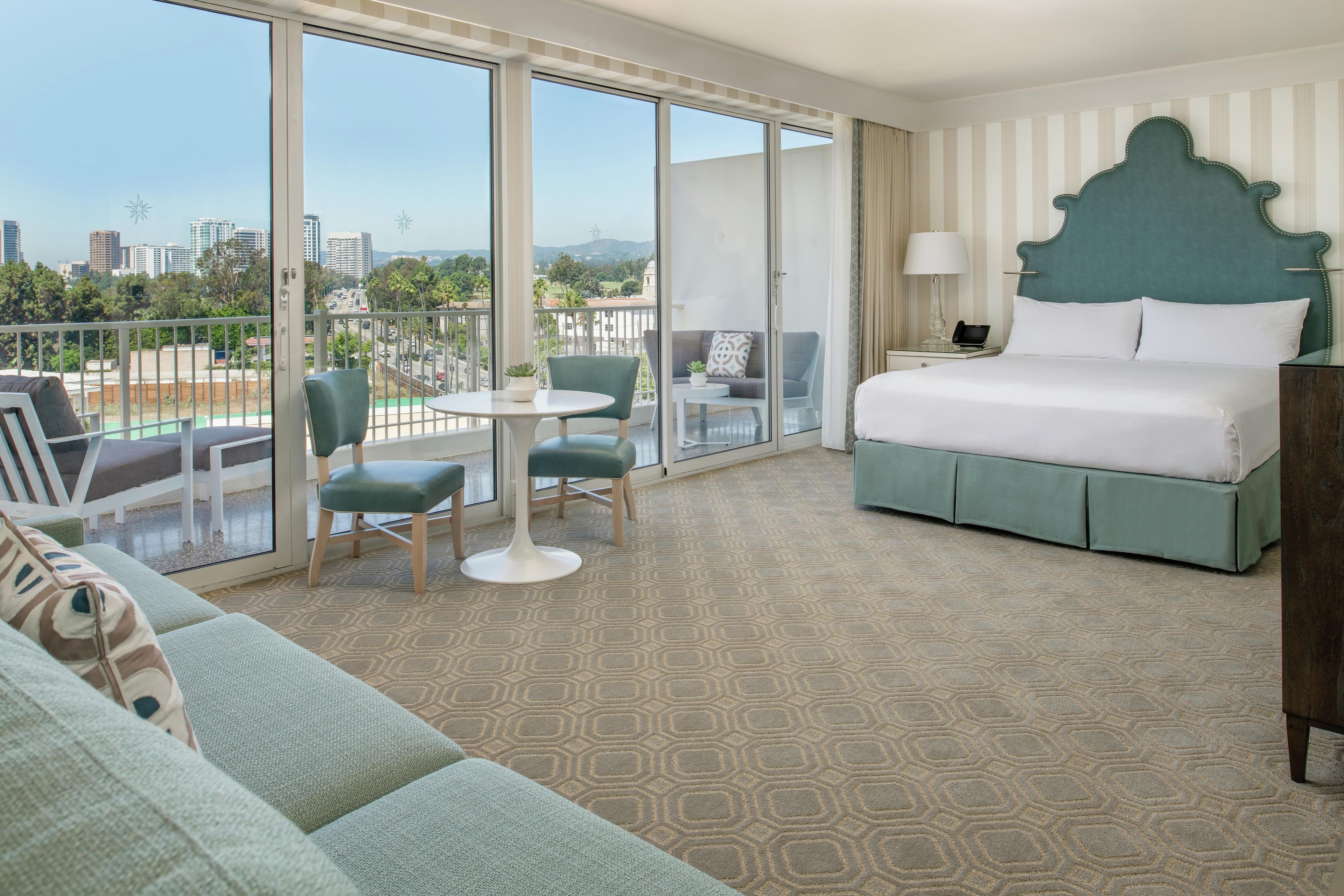 A hotel room with a bed, a balcony with a view of the city, a dining area with a table and chairs, and a sofa.