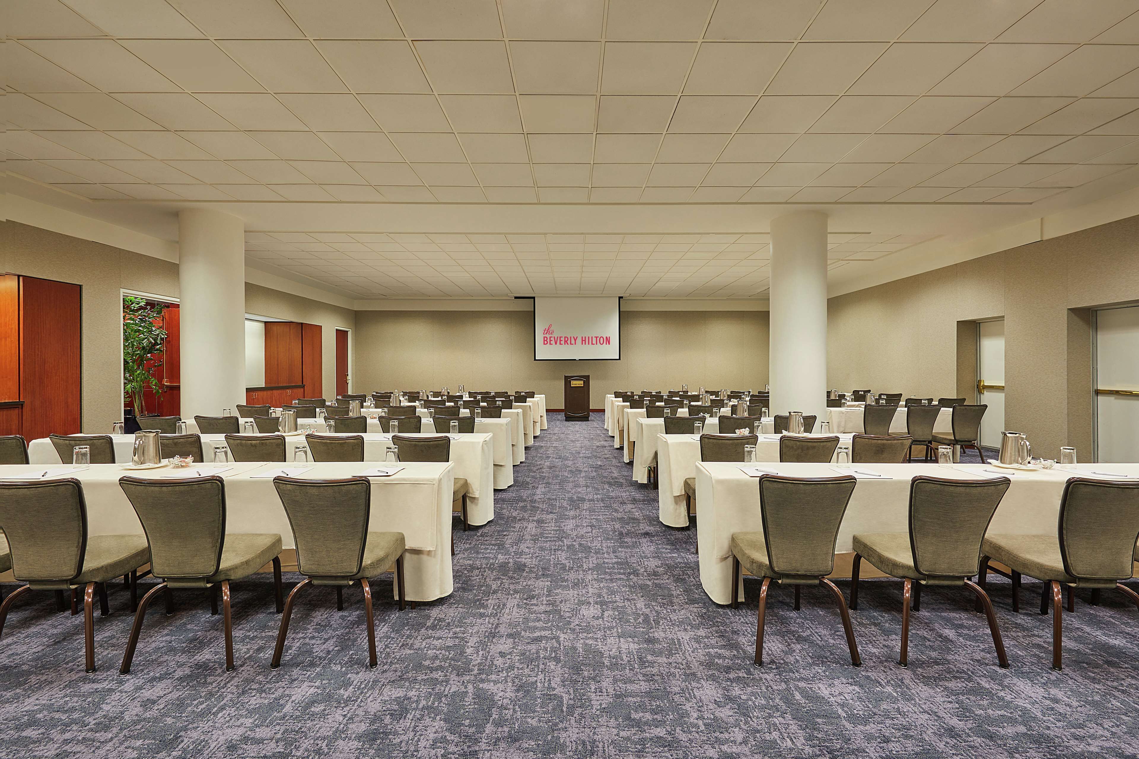 A large conference room with rows of tables and chairs, a carpeted floor, and a projection screen at the front.