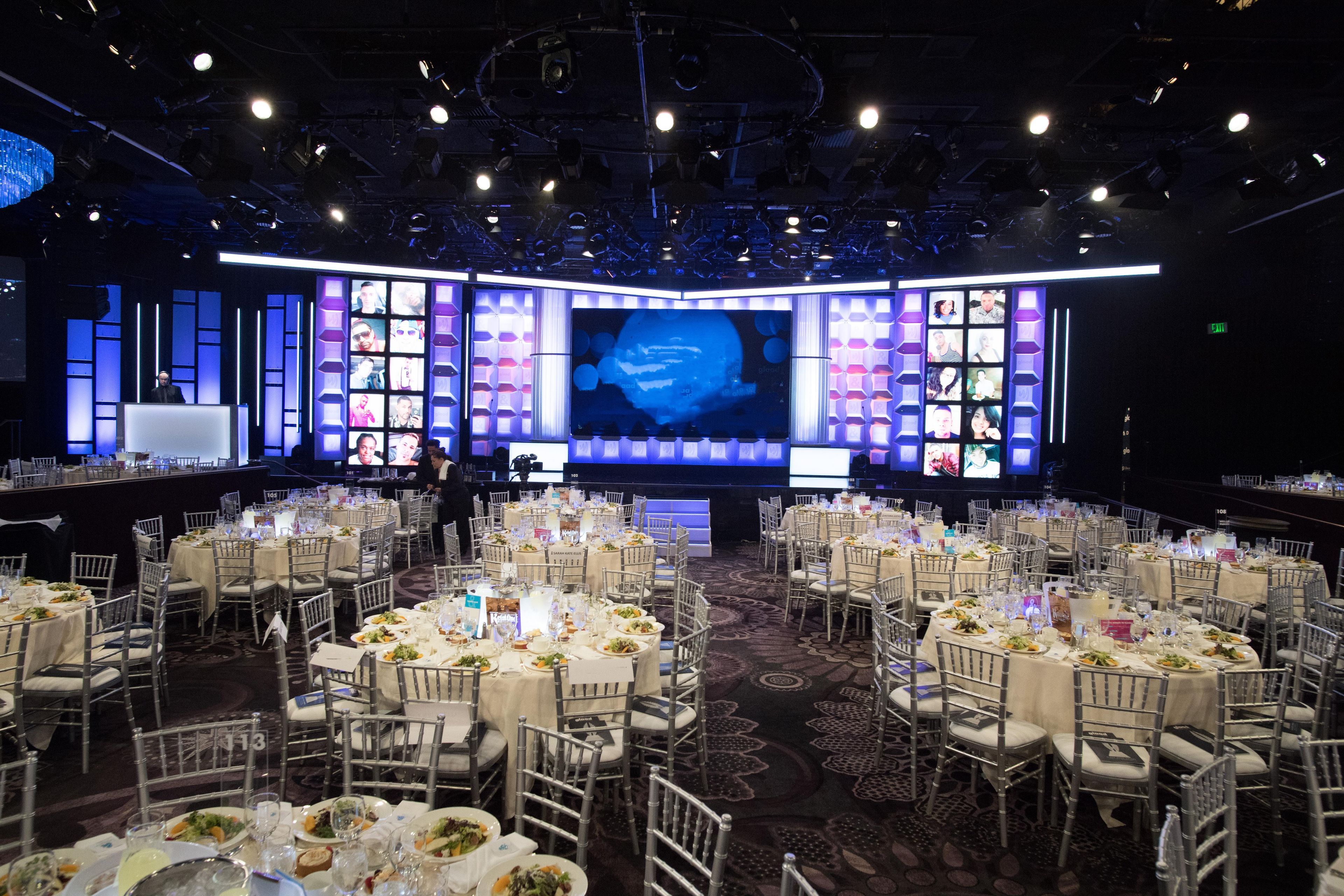 A banquet hall with round tables set for a formal event, a stage with a large screen displaying images, and a dark ceiling with stage lighting.
