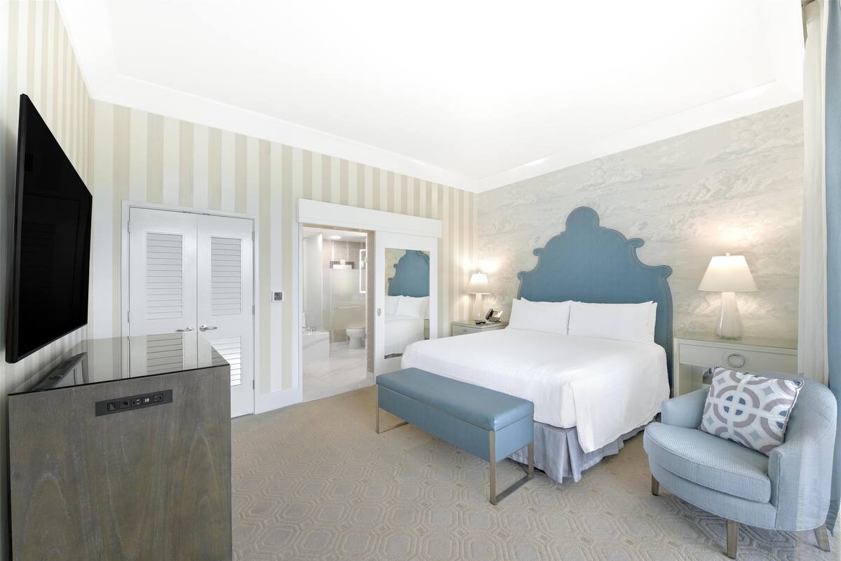 Une chambre d’hôtel avec un lit, une chaise, un banc, une télévision et une salle de bain visible à travers une porte ouverte.