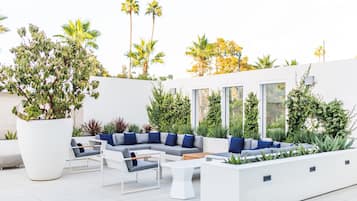 Un área de estar al aire libre moderna con mobiliario blanco, plantas en macetas grandes y vistas al cielo.