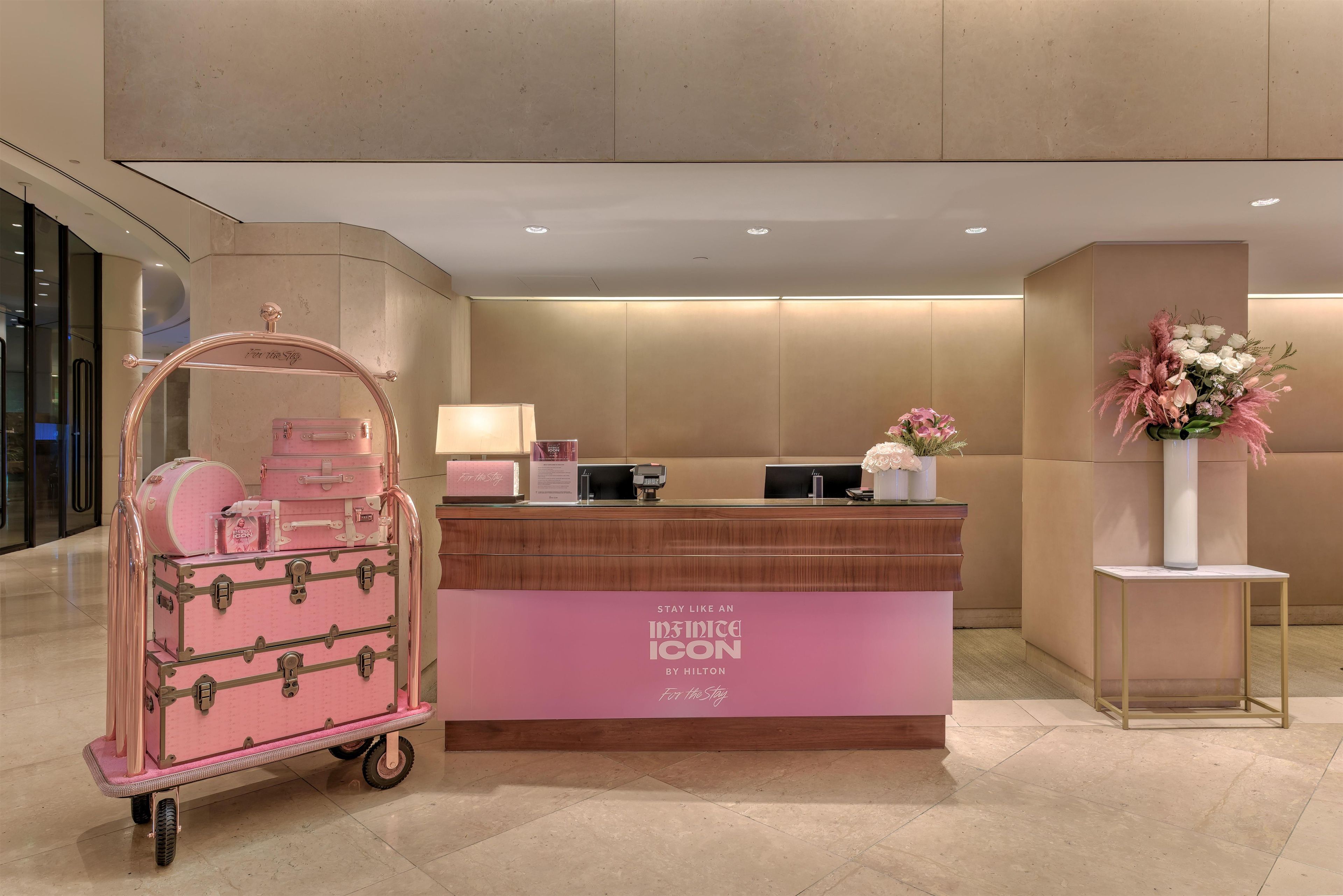 A hotel reception desk with a pink luggage cart, a floral arrangement, and a sign reading 'Stay Like an ICON By Hilton For the Stay'.