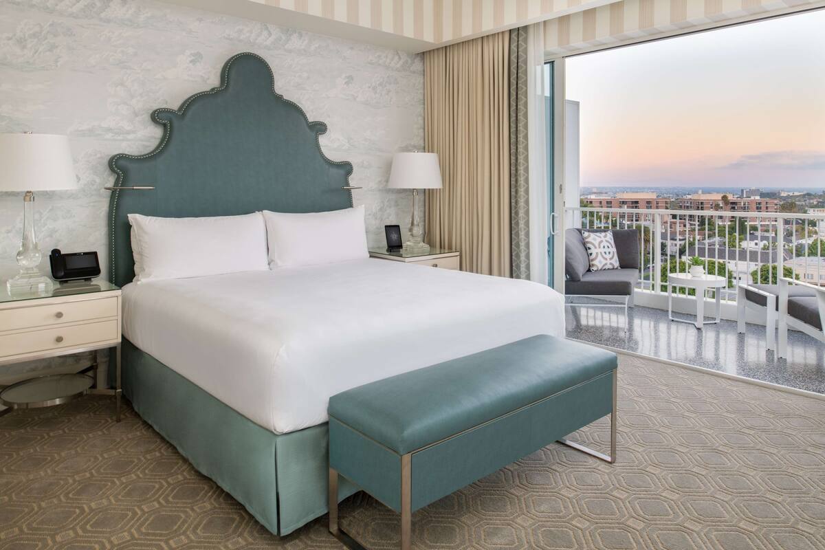 Une chambre d’hôtel avec un grand lit, une banquette, deux tables de chevet et une vue sur la ville depuis un balcon.