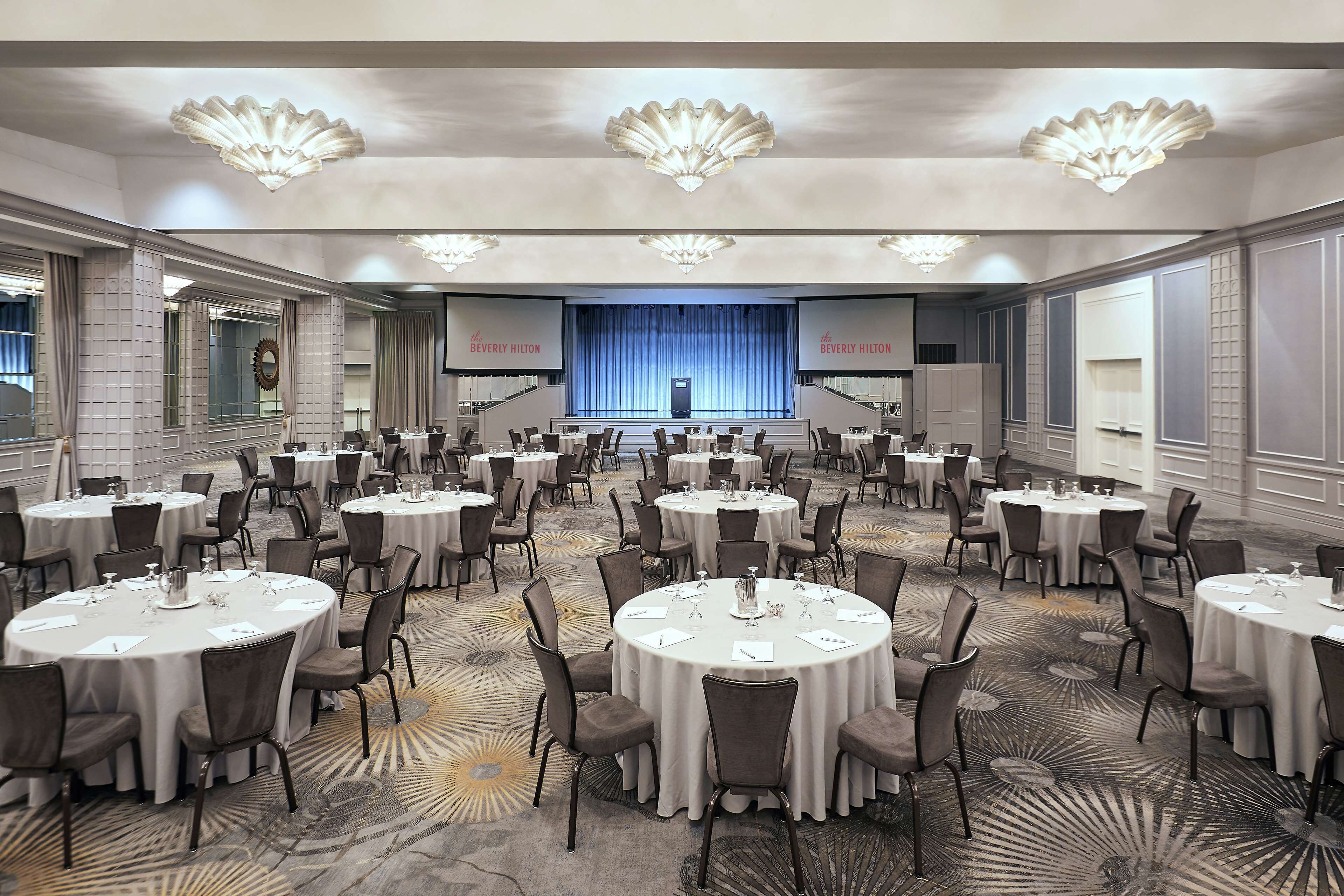 Un salón de conferencias amplio con mesas y sillas redondas, un escenario con cortina azul, y el logotipo del Beverly Hilton en las paredes.
