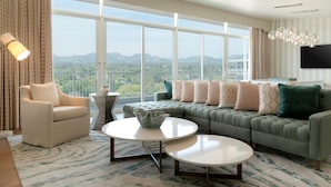 A modern living room with a green sofa, beige armchair, and two white coffee tables. A large window offers a view of the mountains. - The Beverly Hilton (Beverly Hills)