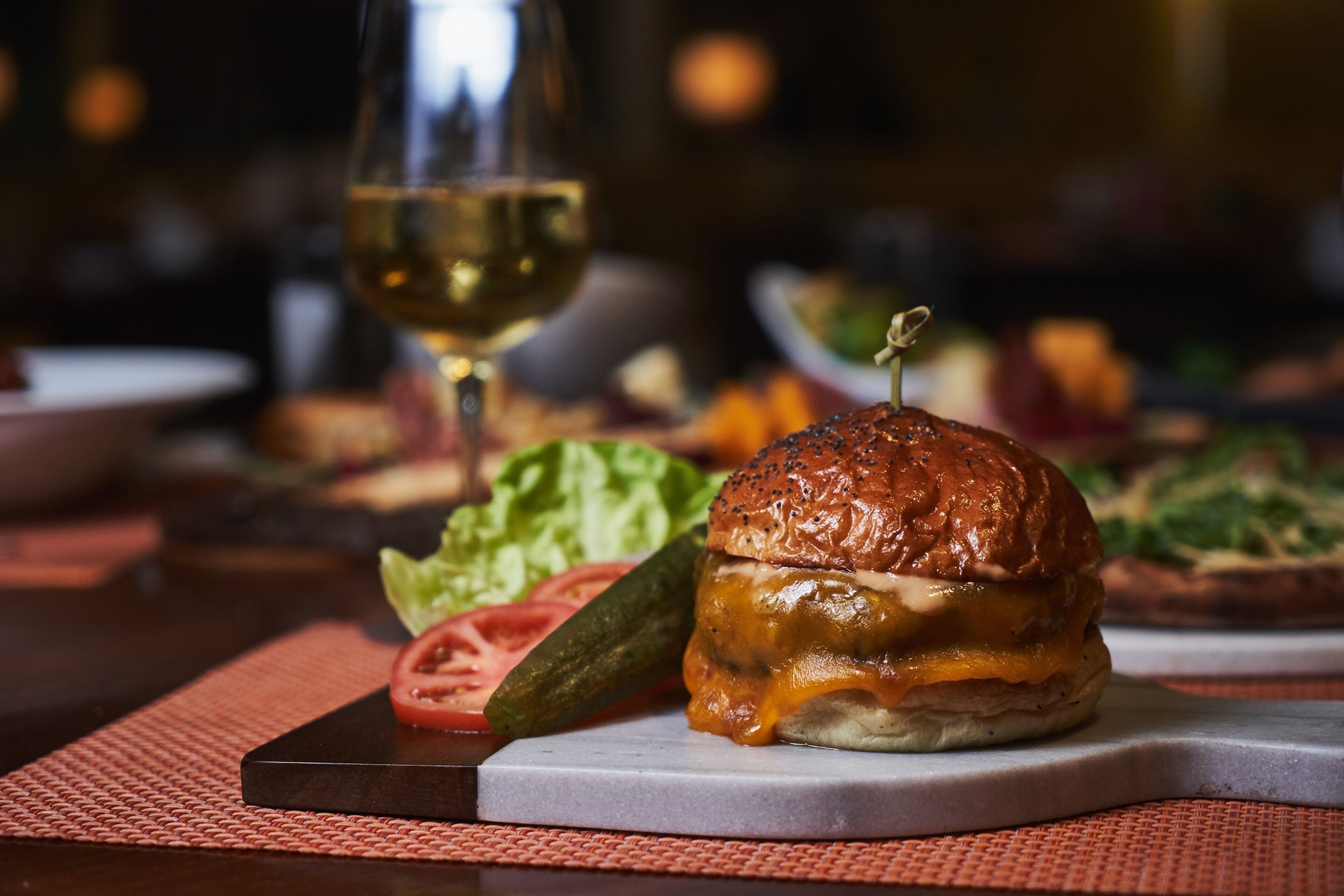 A burger with a sesame seed bun, cheese, and a garnish on top, placed on a white plate with a red placemat underneath.