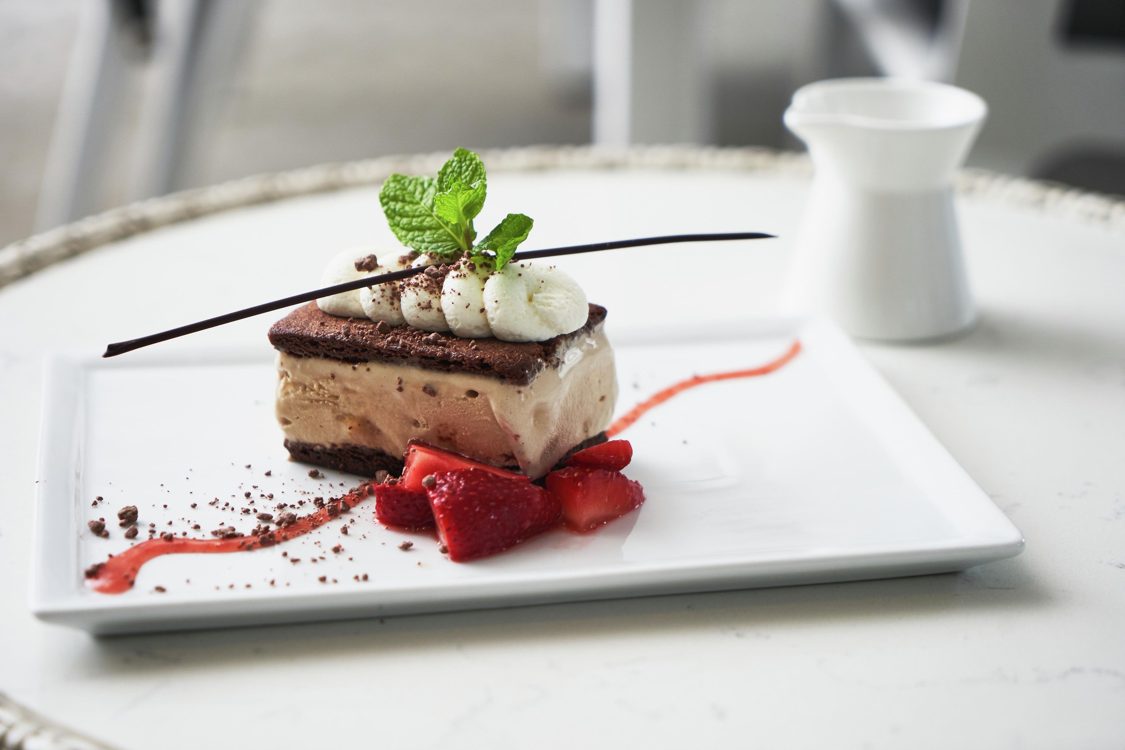 A dessert on a white plate with a chocolate and vanilla layer, garnished with mint leaves and strawberries.