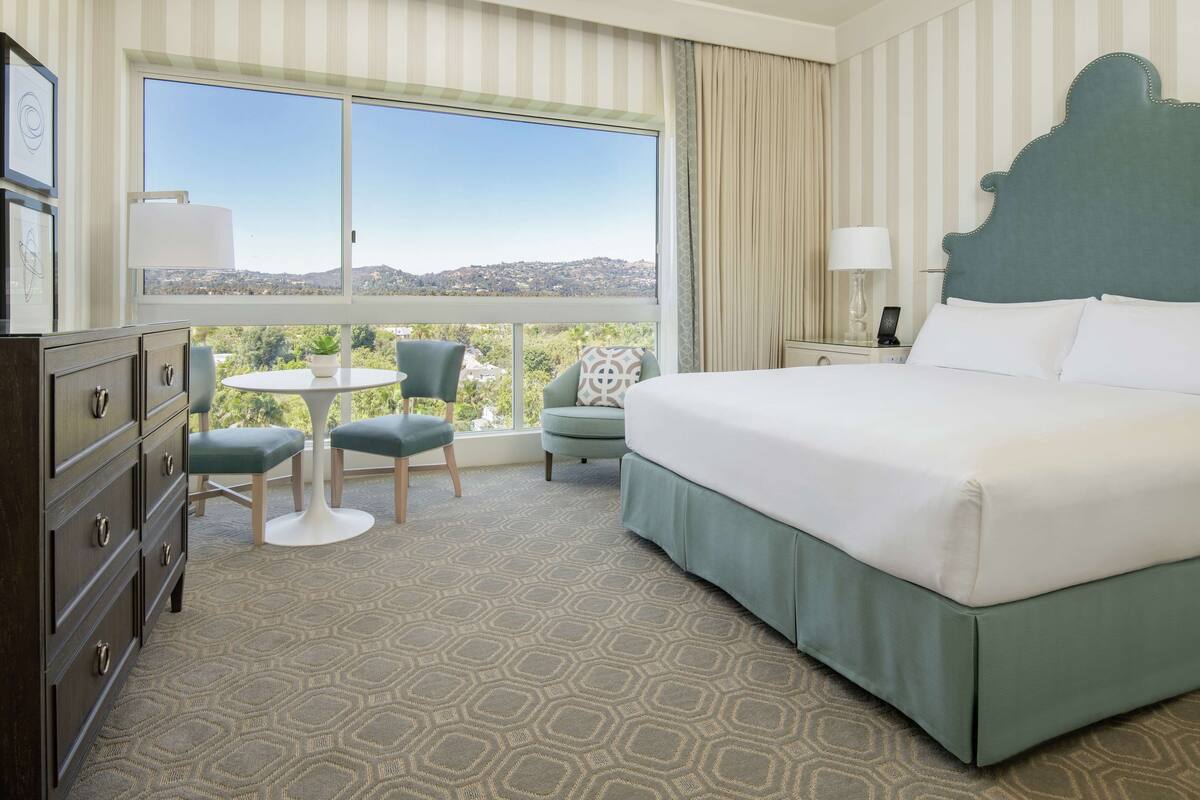 Une chambre d’hôtel avec un grand lit, un bureau, une chaise et une vue sur la ville grâce à de grandes fenêtres.