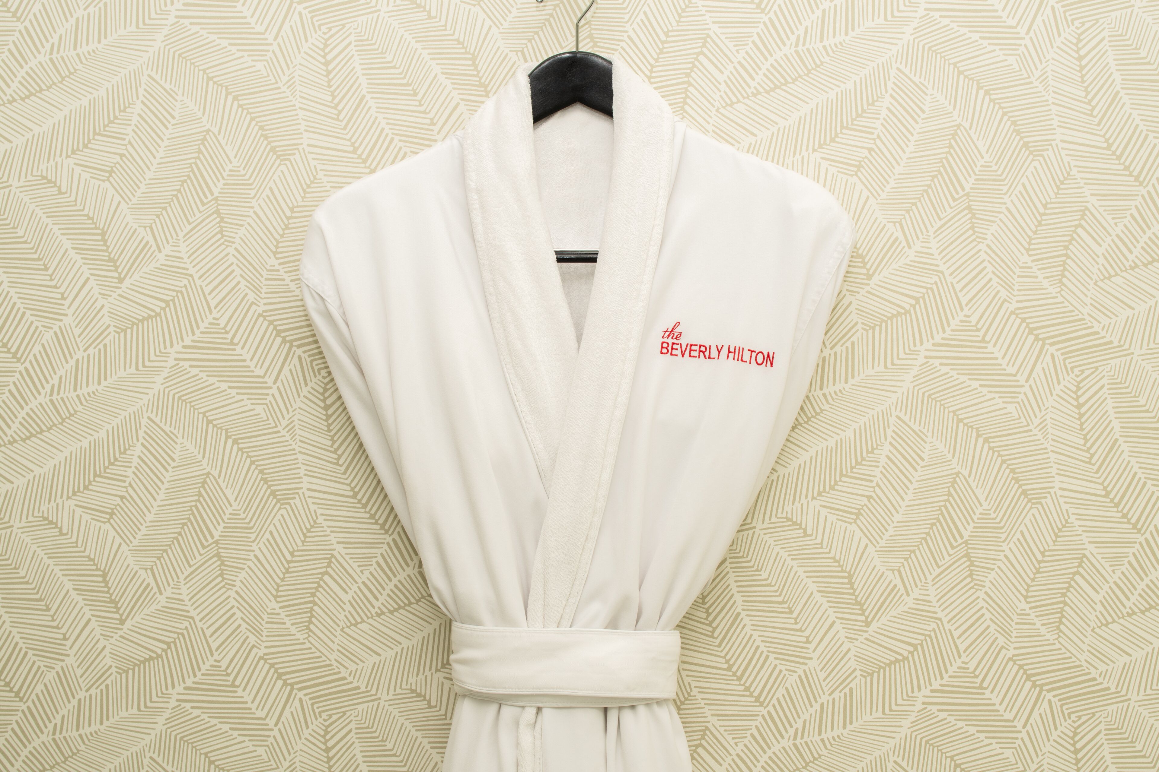 Um roupão branco com o logotipo do Beverly Hilton pendurado em um cabide preto contra uma parede estampada.
