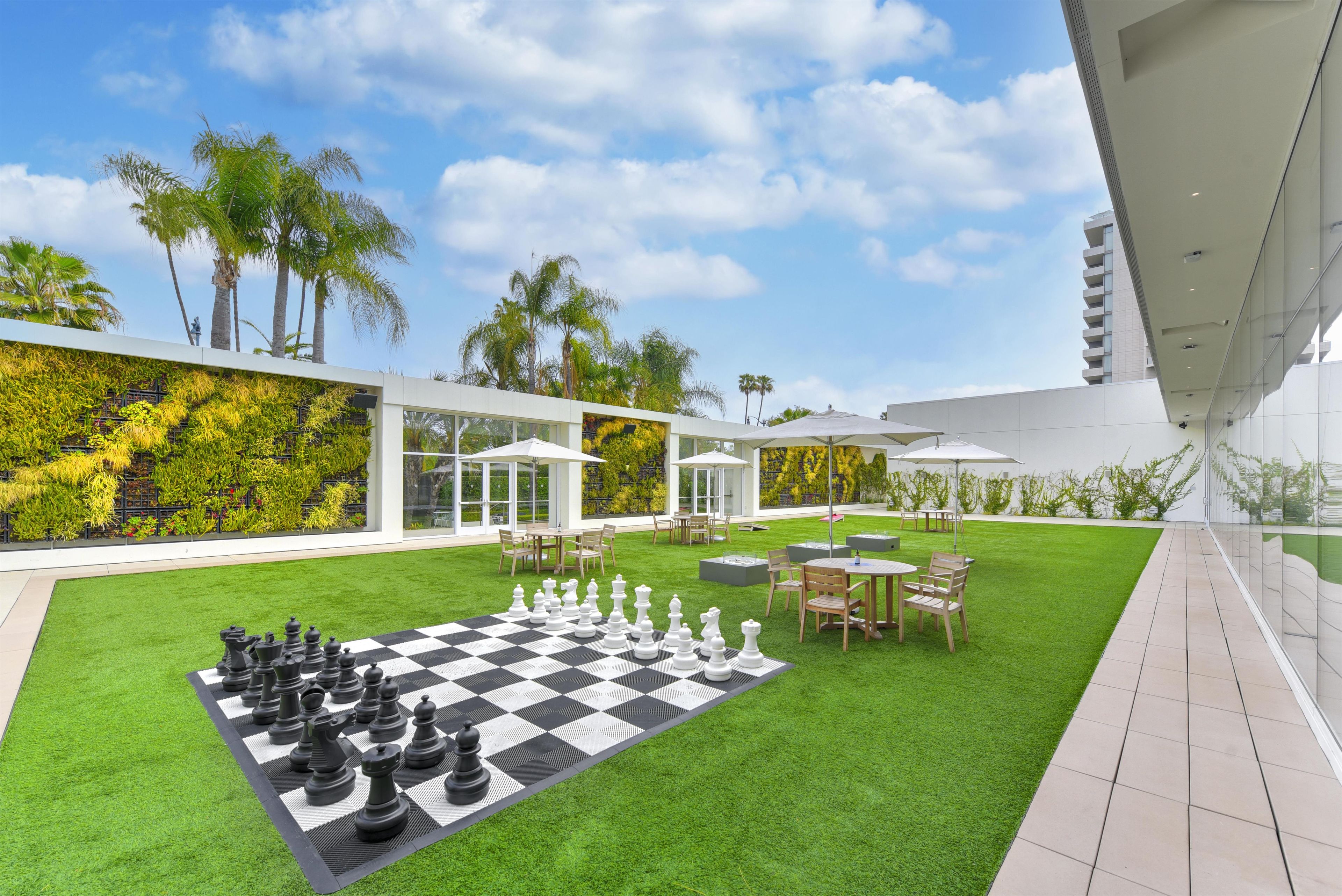 A spacious outdoor area with a chessboard set up on artificial grass, surrounded by modern outdoor furniture and palm trees.