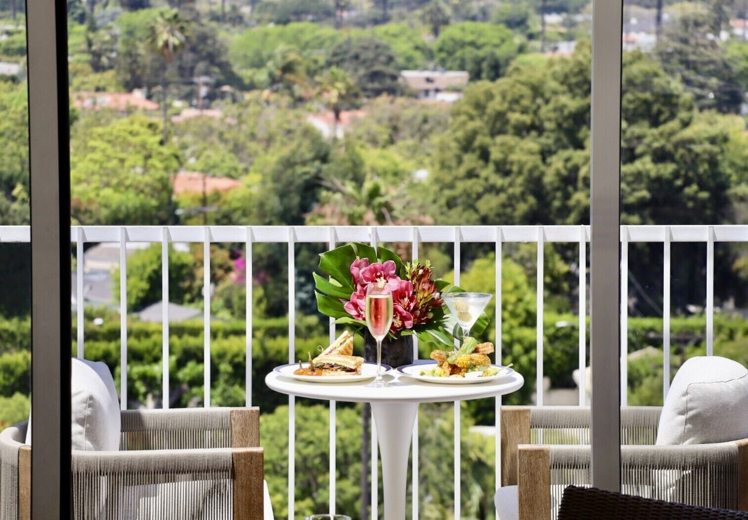 A balcony with a table set for two, featuring a meal, a drink, and a floral arrangement.