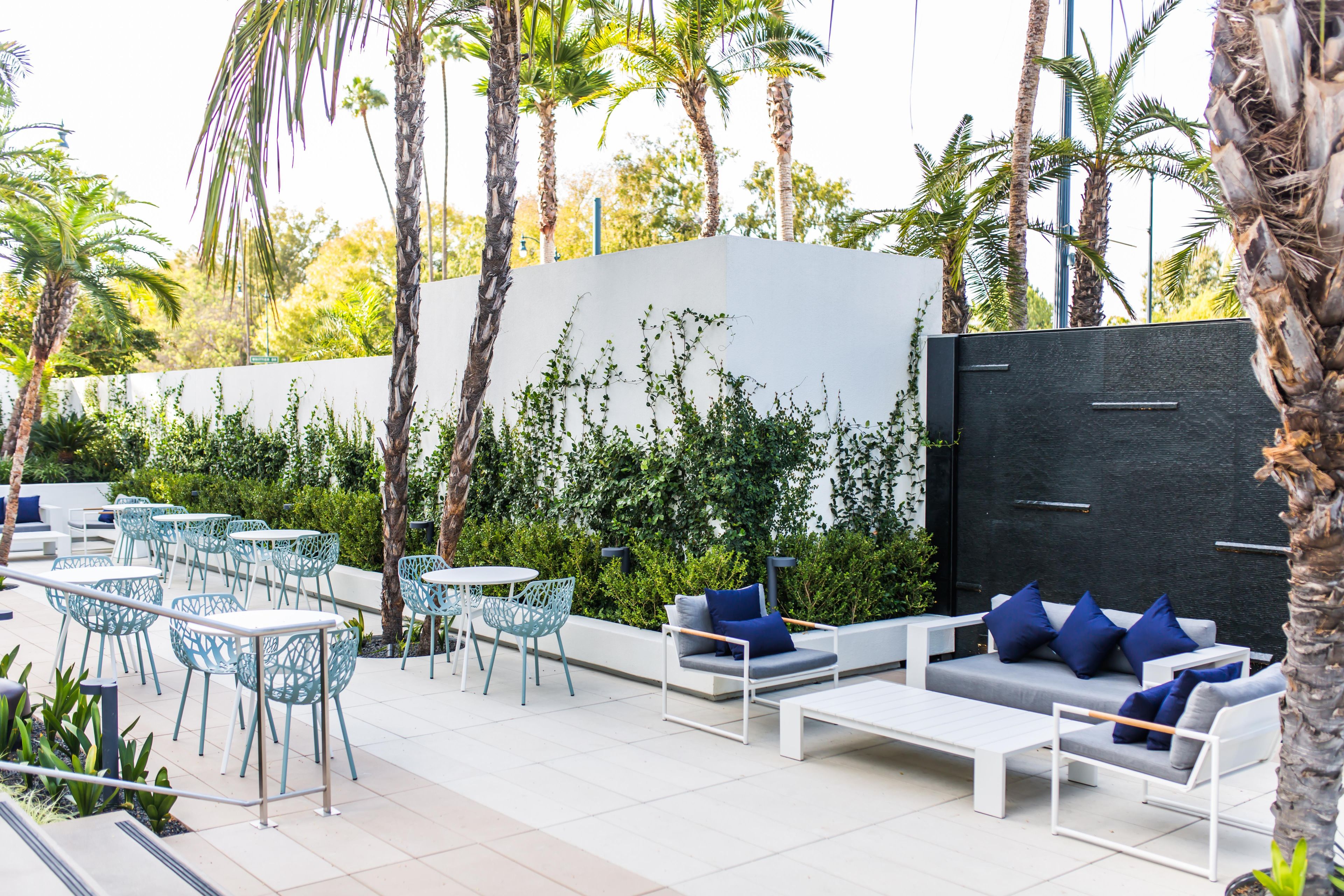 An outdoor seating area with white sofas and blue cushions, surrounded by palm trees and a white wall with greenery.