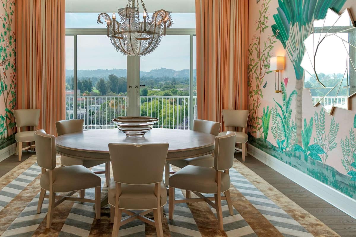 Un espace repas comprenant une table ronde et six chaises, un lustre et une vue sur la verdure grâce à de grandes fenêtres.