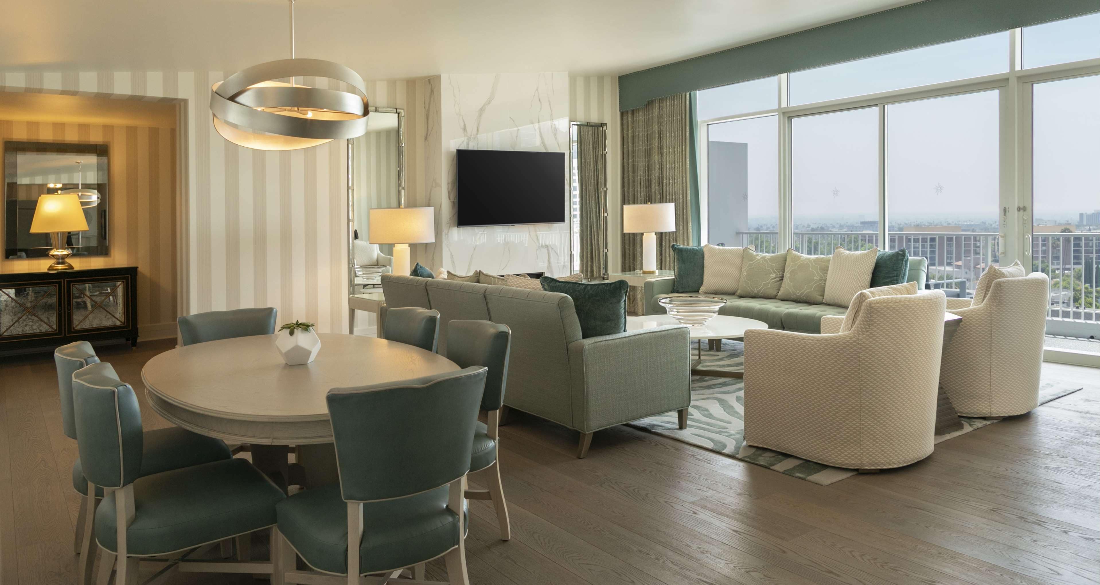 A modern hotel room with a large window, a flat-screen TV, a dining area with a round table and chairs, and a seating area with sofas and armchairs.