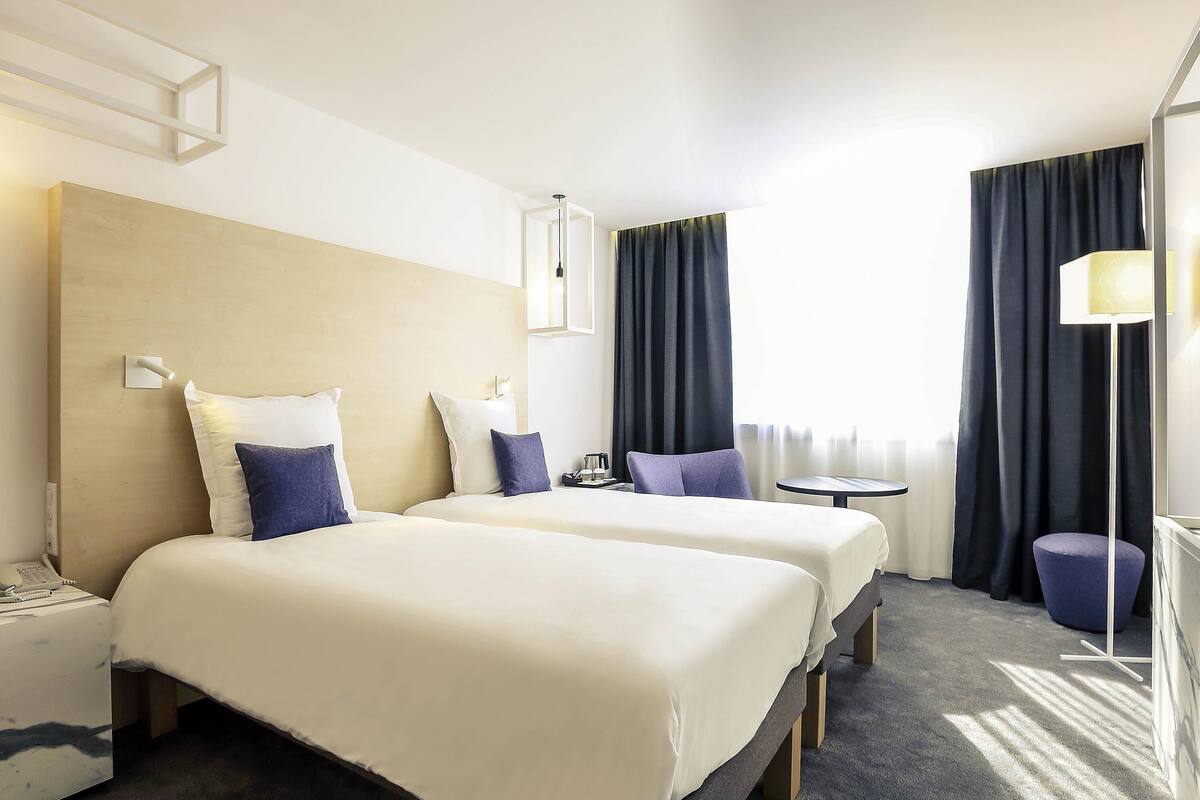 Habitación de hotel con dos camas, una mesita de noche con lámpara, una mesa redonda pequeña y una silla.