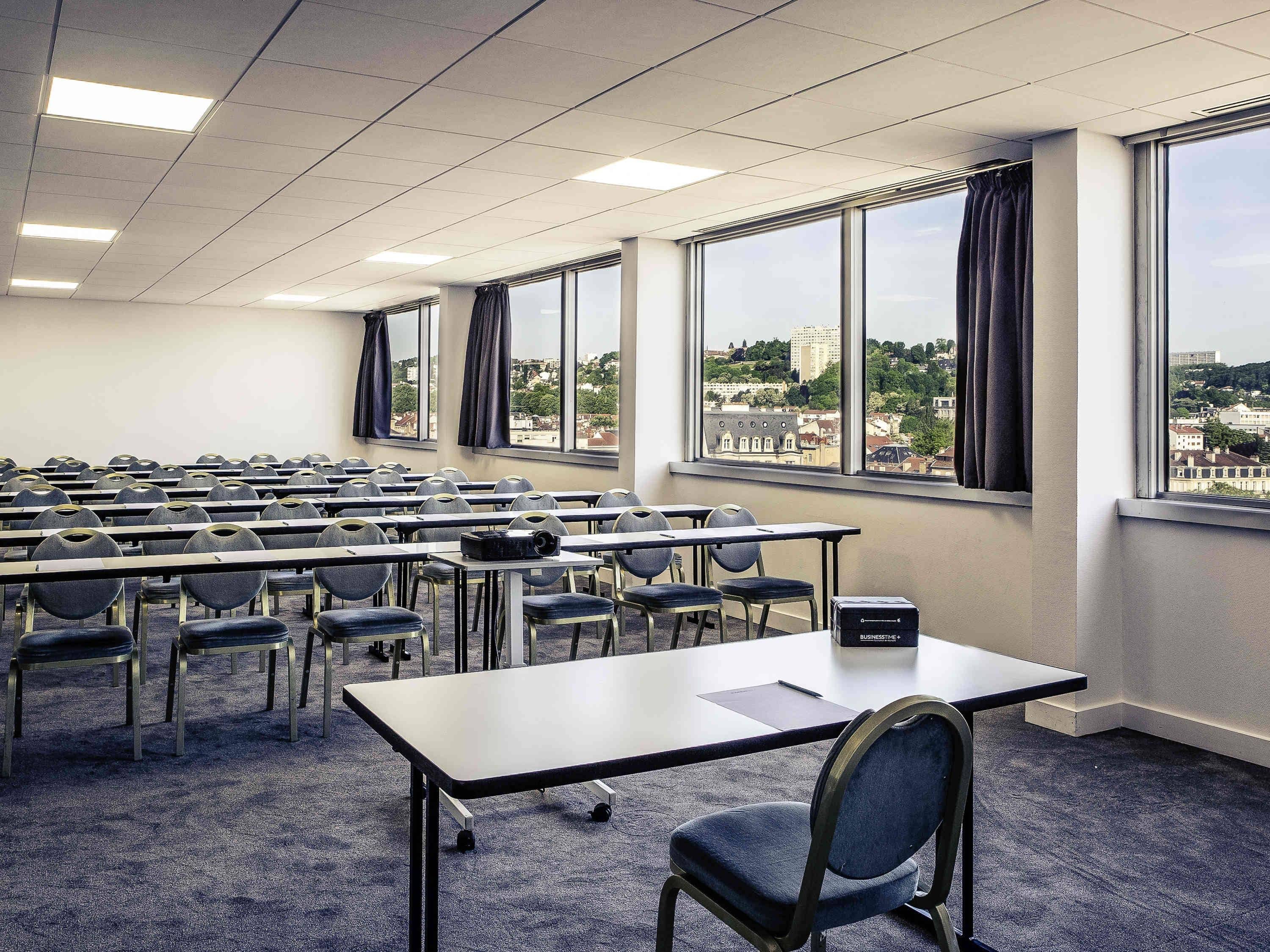 Un salón de conferencias con filas de mesas y sillas, ventanas amplias con cortinas y vistas a la ciudad.