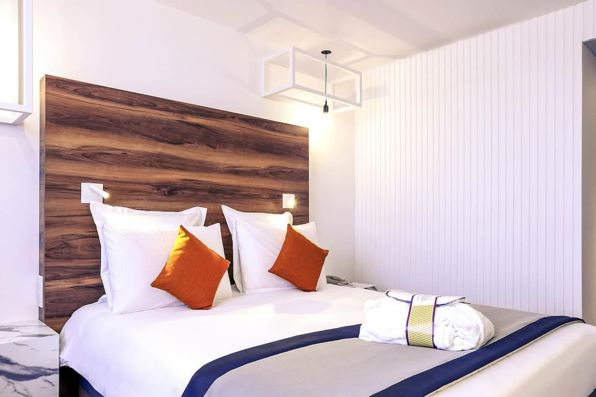 Quarto de hotel com cabeceira de madeira, roupa de cama branca, almofadas laranja e um toque de azul, além de uma mesa de cabeceira com abajur.
