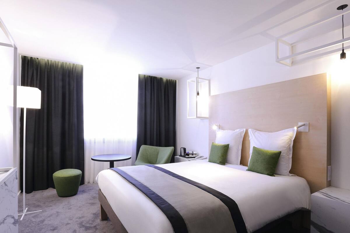 Una habitación de hotel con una cama grande, una mesita redonda pequeña, una silla verde y una mesita de noche.