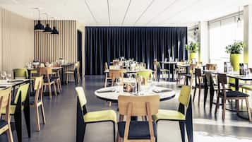 Un restaurante moderno con mesas y sillas redondas, una pared de acento de madera y un gran ventanal con plantas.