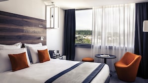 Quarto de hotel com uma cama grande, uma mesa pequena, uma cadeira e vista para a cidade pela janela.