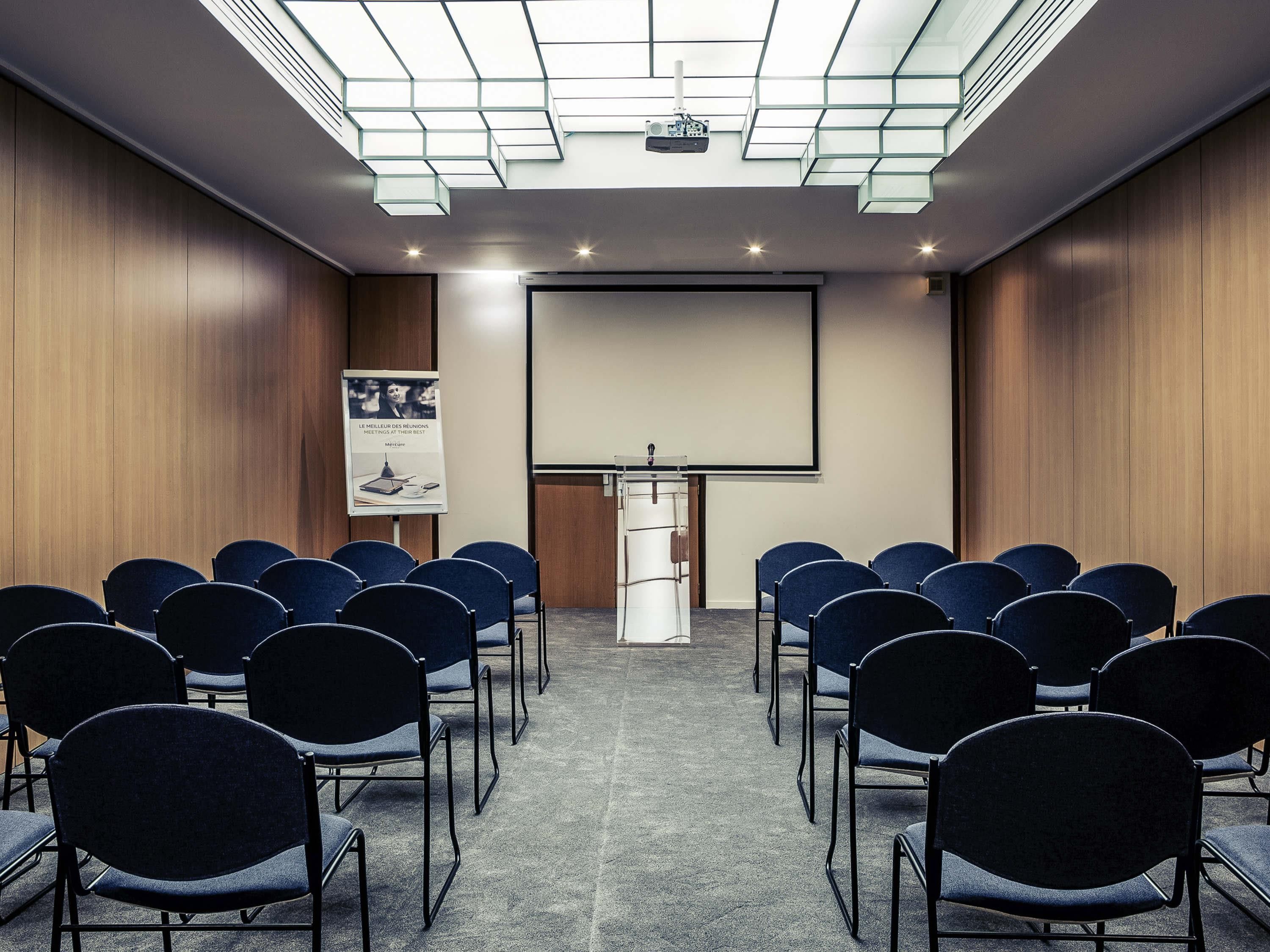 A conference room with rows of chairs, a podium, a projector screen, and a poster on the wall.