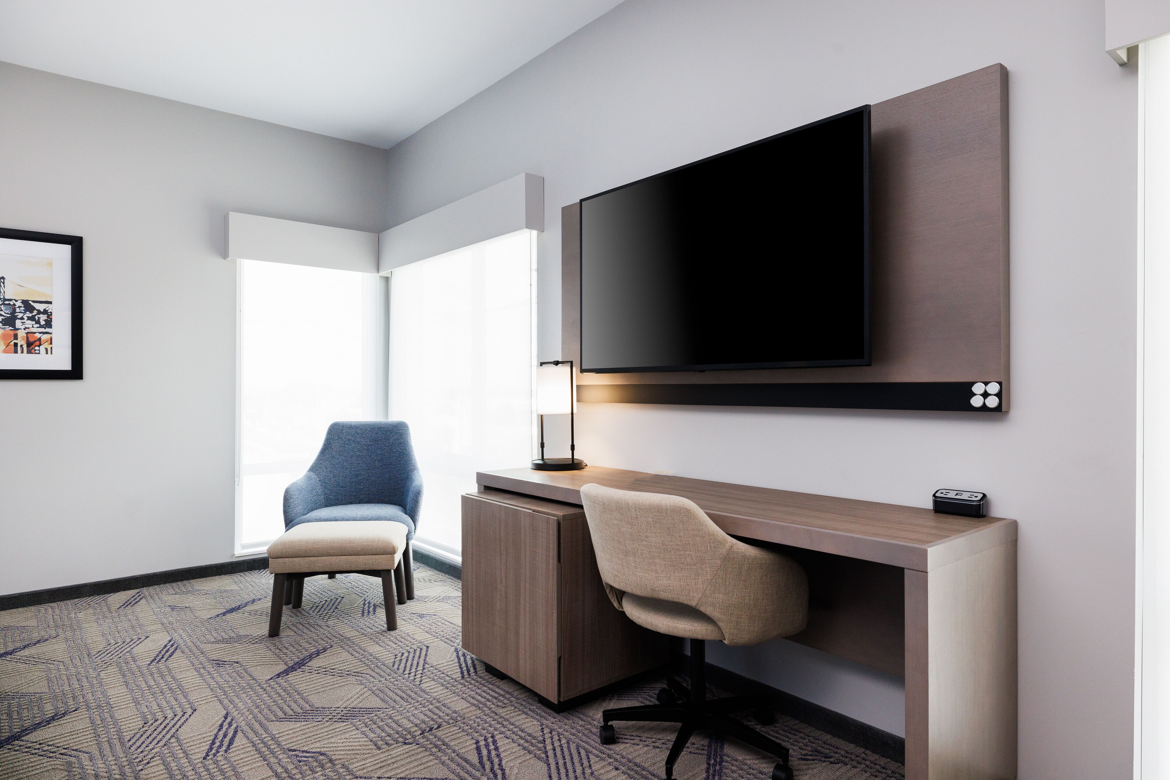 A modern hotel room with a large flat-screen TV mounted on the wall, a wooden desk, a comfortable armchair, and a patterned carpet.