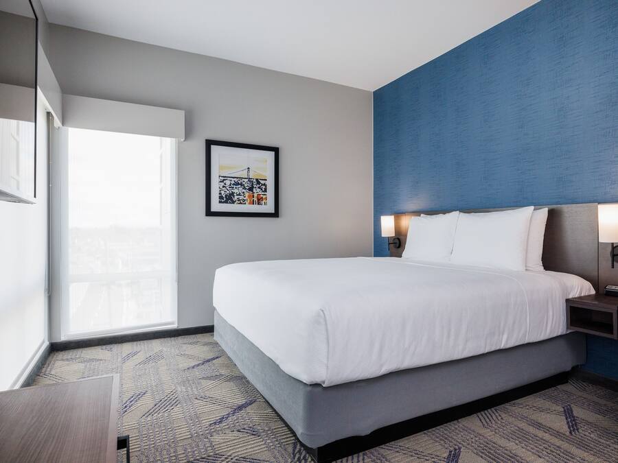 Quarto de hotel com uma cama grande, mesa de cabeceira, um quadro na parede e janela com vista.