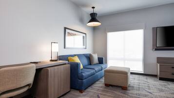 A modern living room with a blue sofa, a small ottoman, a desk with a lamp, a TV, and a framed picture on the wall.