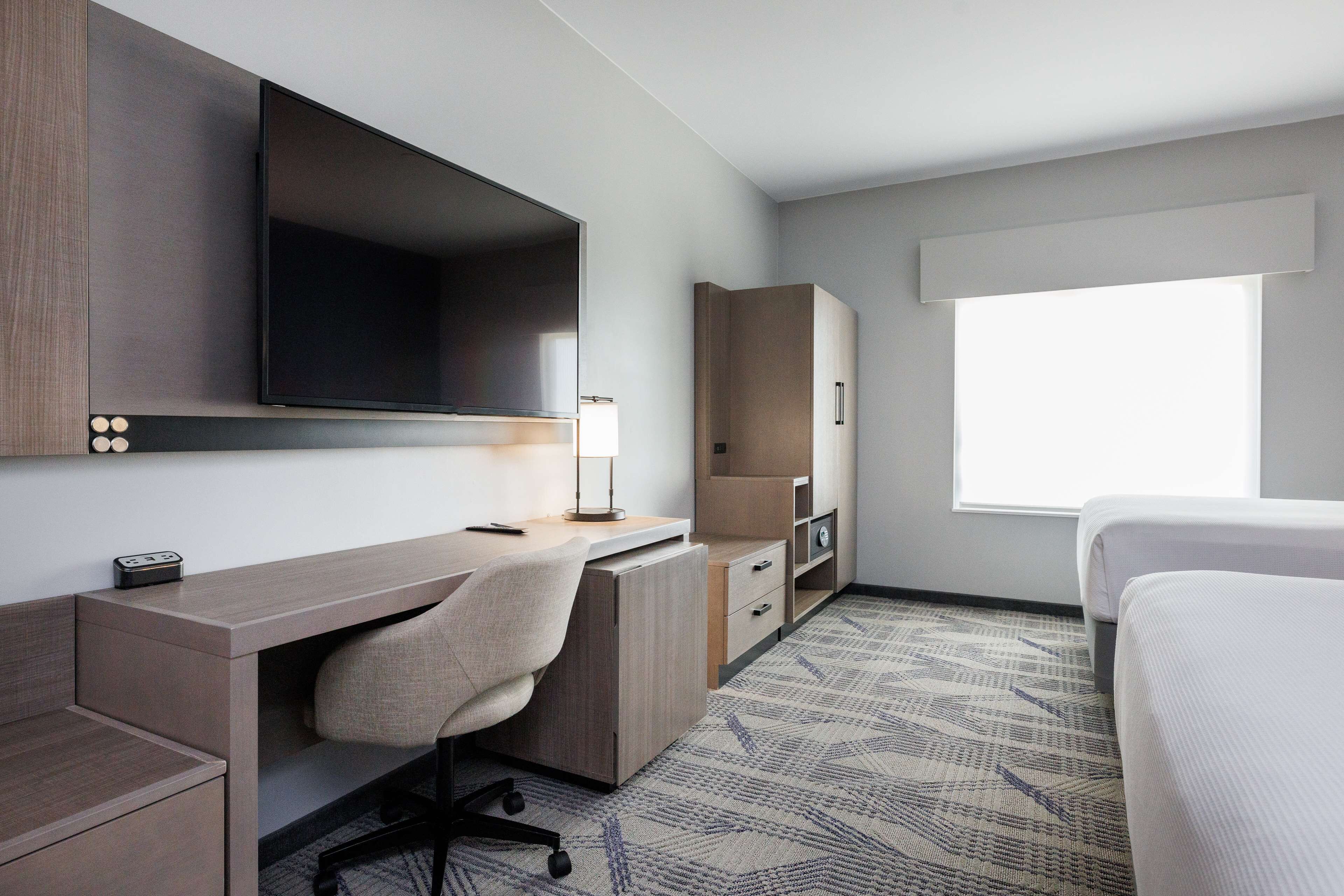 Une chambre d’hôtel comprenant un lit, un bureau avec une chaise, un téléviseur à écran plat, une lampe et une fenêtre laissant entrer la lumière du jour.