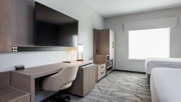 Habitación de hotel con cama, escritorio con silla, televisor de pantalla plana, lámpara y ventana con luz natural.