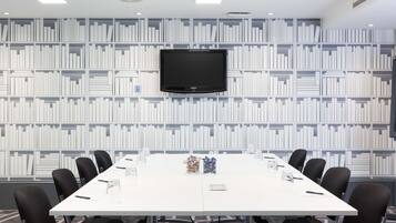 A conference room with a white table, black chairs, a Samsung TV mounted on the wall, and a bookshelf in the background.