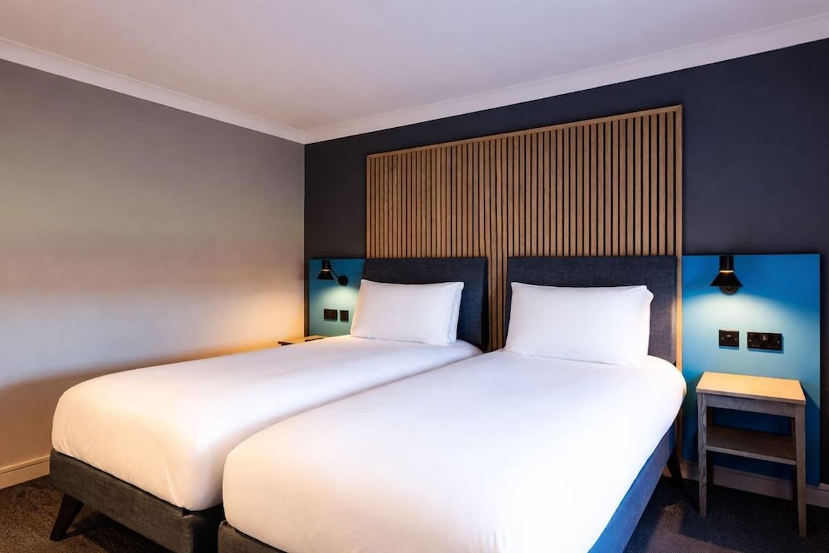 A hotel room with two beds, a wooden headboard, bedside lamps, and a small table.