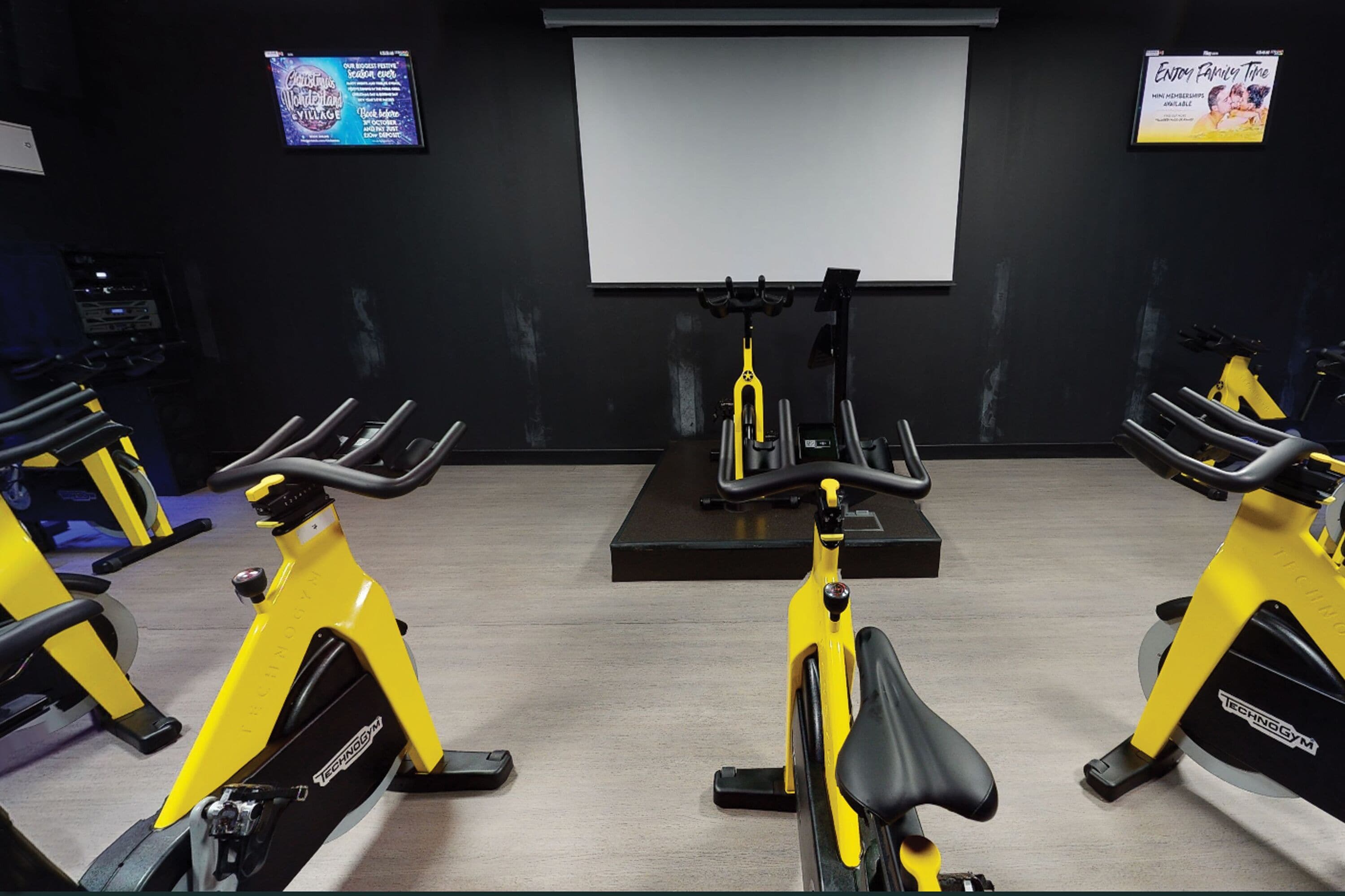 Uma academia com bicicletas ergométricas, uma tela de projeção e uma TV fixada na parede.