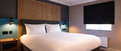 Una habitación de hotel con una cama grande, cabecera de madera, dos mesitas de noche con lámparas, una ventana con persiana y un termostato en la pared.