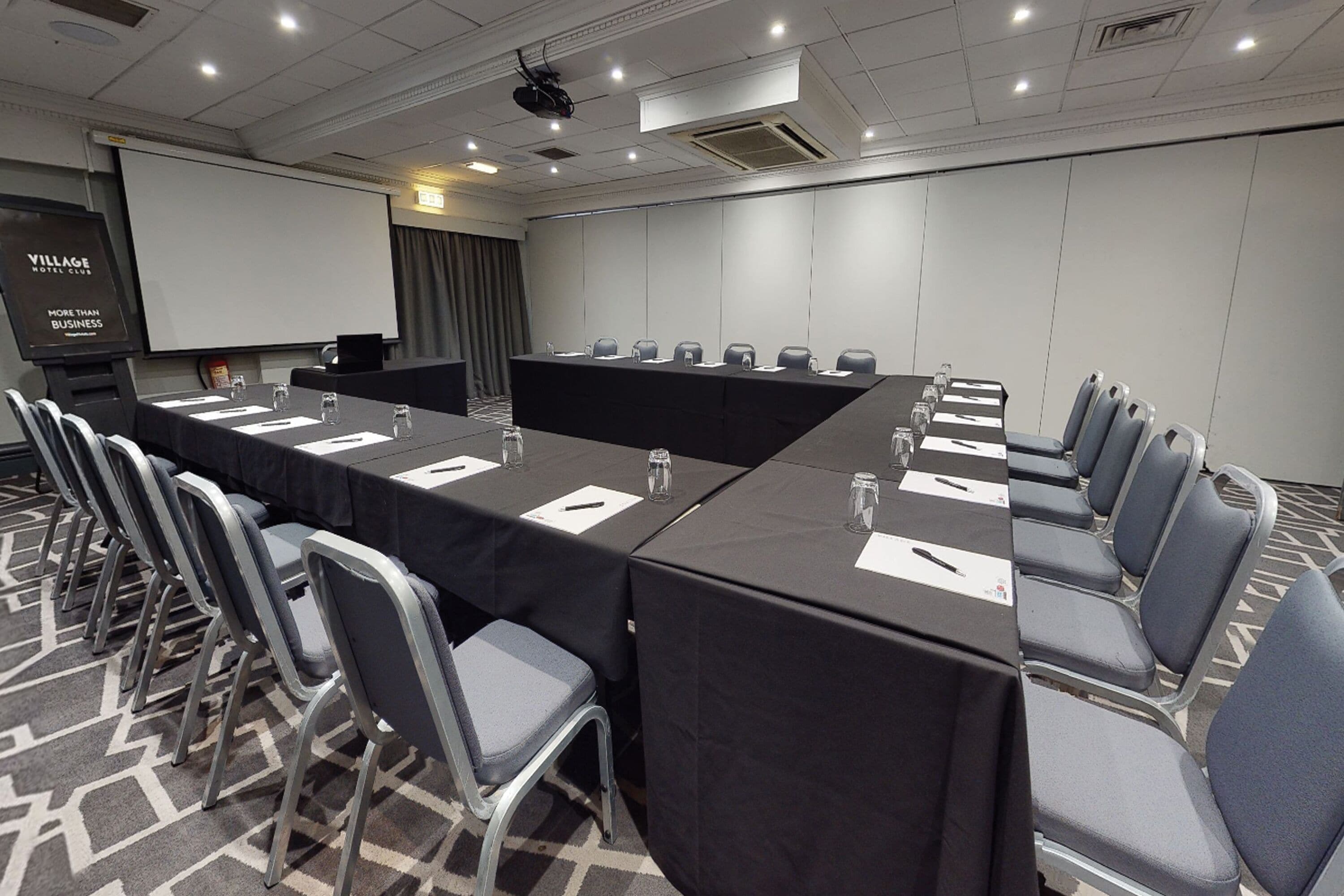 A conference room with a long table, chairs, a projector screen, and a screen displaying "VILLAGE HOTEL CLUB MORE THAN BUSINESS".