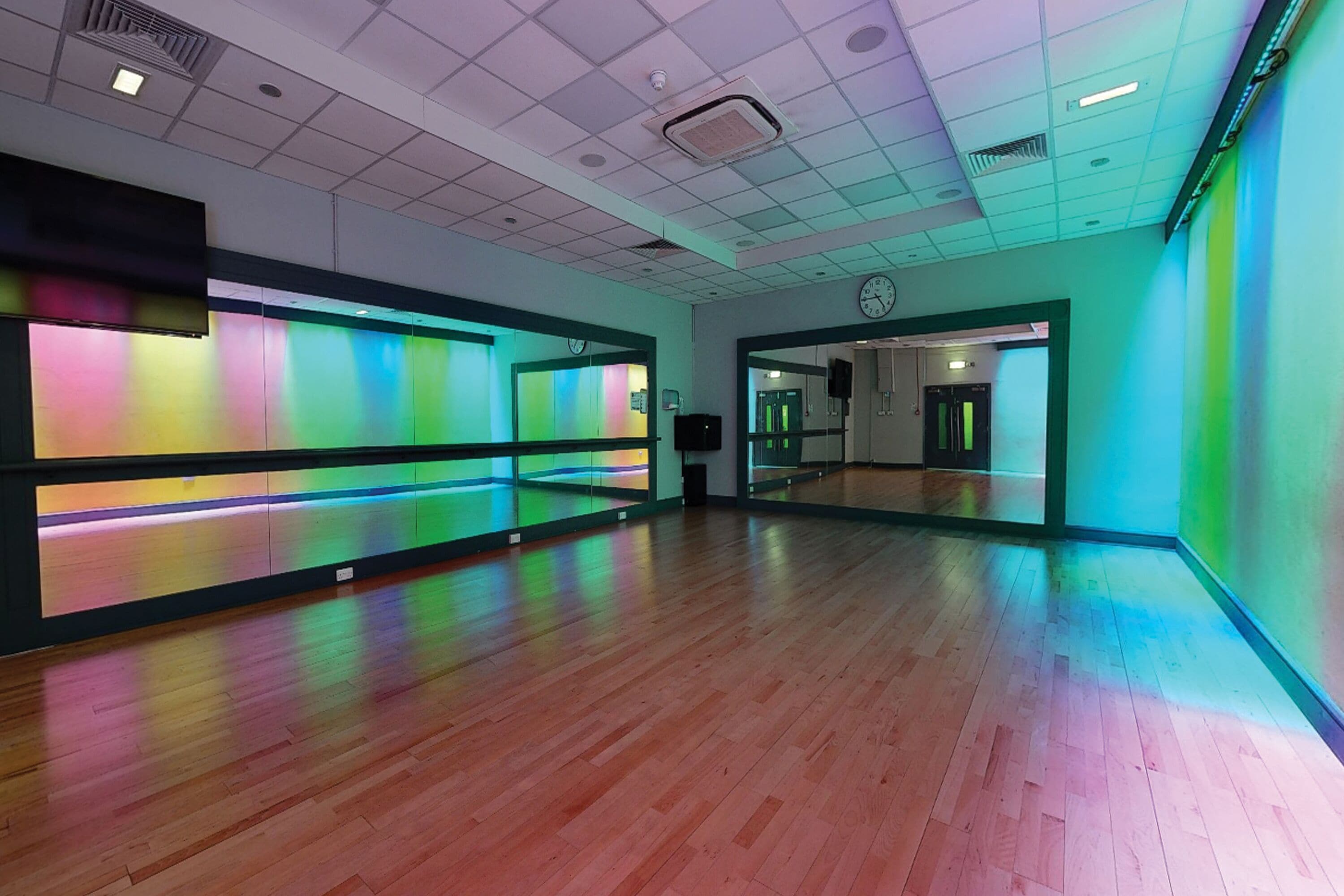 A spacious room with a large mirror, wooden flooring, and a colorful light strip on the wall.