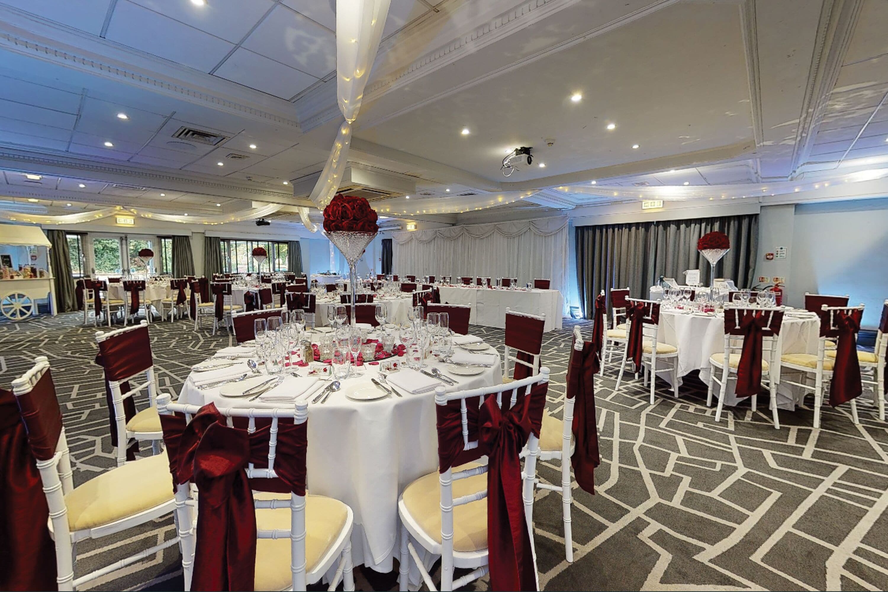 A banquet hall with round tables set for dining, white chairs with red sashes, and a chandelier centerpiece.