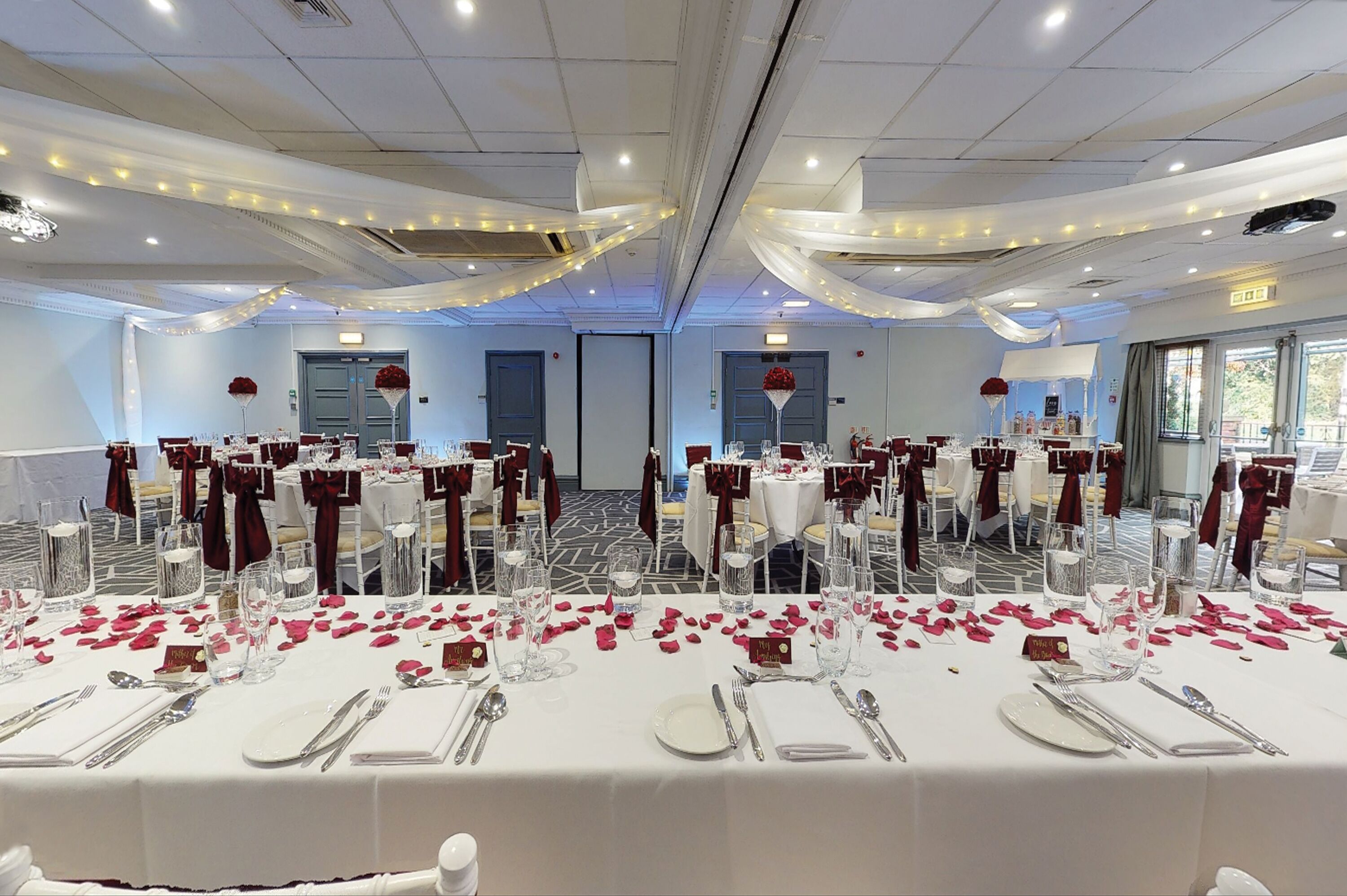 Een feestzaal met ronde tafels gedekt voor een diner, bedekt met witte en rode stoffen, en versierd met rozenblaadjes.