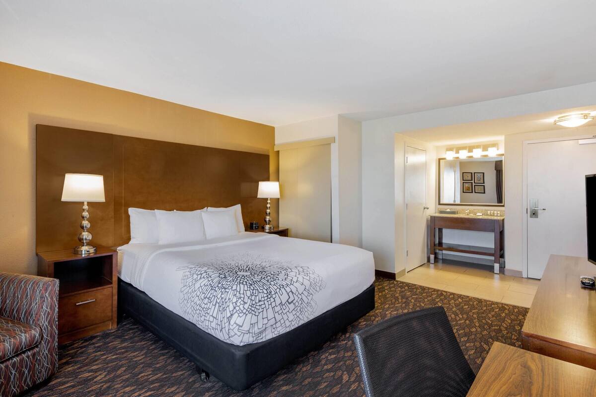 Hotelkamer met een groot bed, twee nachtkastjes met lampen, een houten hoofdbord, een zithoekje met een stoel, een bureau en een badkamer met spiegel en wastafel.