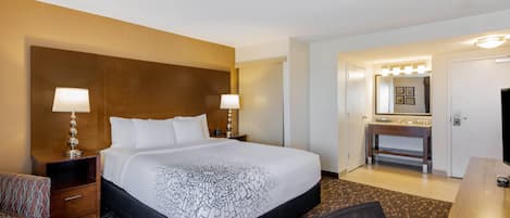 Una habitación de hotel con una cama grande, dos mesitas de noche con lámparas, un cabecero de madera, una pequeña zona de estar con una silla, un escritorio y un baño con espejo y lavamanos.